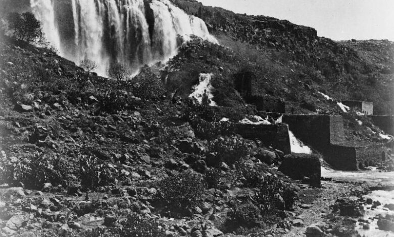 A broad waterfall cascades over a rocky cliff into a stream below, with stone structures along the bank and rugged terrain surrounding the watercourse.
