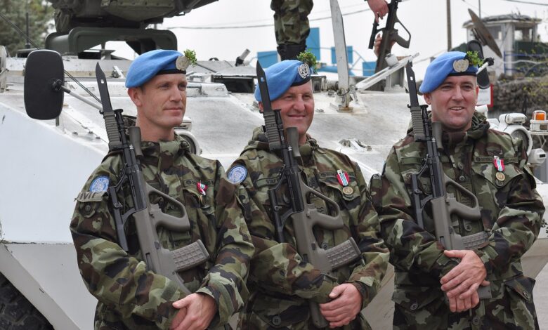 Three UN peacekeepers wearing blue berets stand in front of a white armored vehicle, holding rifles and posing for a medal ceremony on the Golan Heights. The setting appears to be a UNDOF base area.