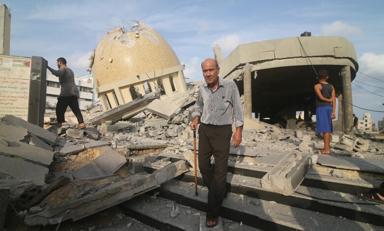 Several Palestinians walk among heavy rubble with a large, collapsed mosque dome and damaged buildings in the background on a clear day.
