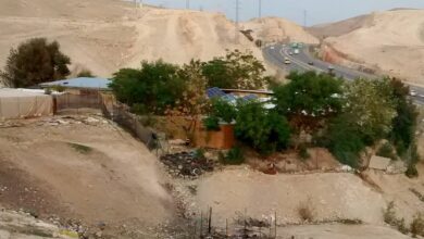 A cluster of simple school buildings in a desert landscape at Khan al-Ahmar, with trees, tires used in the grounds, and Israeli Highway 1 running behind the site through the arid hills.