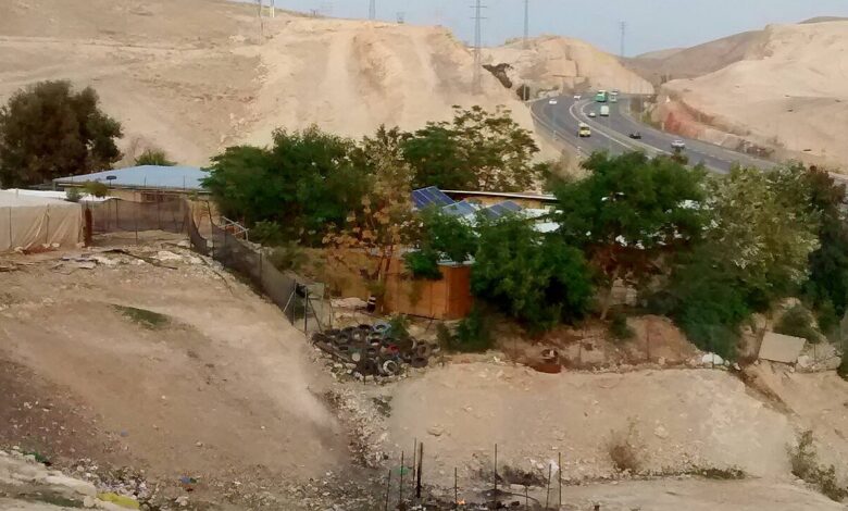 A cluster of simple school buildings in a desert landscape at Khan al-Ahmar, with trees, tires used in the grounds, and Israeli Highway 1 running behind the site through the arid hills.