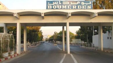 A wide road passing under a white gateway structure bearing the word “BOUMERDES” in Latin and Arabic script, with sidewalks, trees, and streetlights leading into the city.