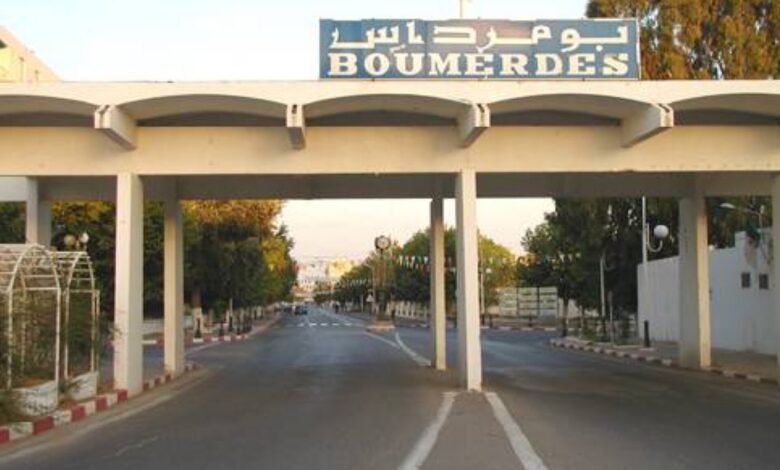 A wide road passing under a white gateway structure bearing the word “BOUMERDES” in Latin and Arabic script, with sidewalks, trees, and streetlights leading into the city.