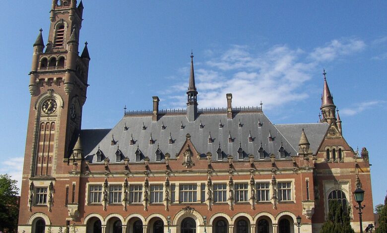 A clear, front-facing view of the Peace Palace in The Hague, showing its central facade, clock tower, arched colonnade, and neatly kept lawn under a blue sky.
