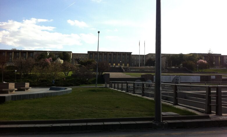 A wide, daylight view of a large government building in Ankara with flagpoles and a landscaped plaza in the foreground, photographed from across a roadway.