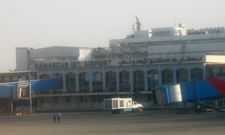 Exterior view of the Damascus International Airport terminal with clear signage reading “DAMASCUS INTL. AIRPORT,” jet bridges, apron equipment, and two people standing on the tarmac.