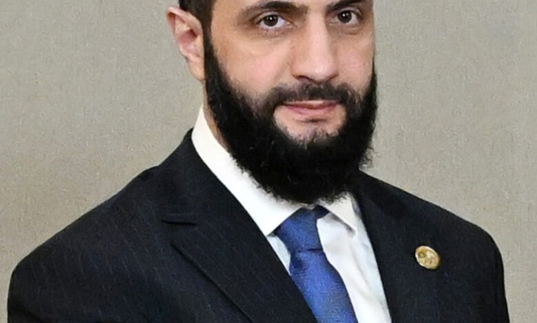 A bearded man in a dark pinstripe suit and tie, facing the camera with a neutral expression against a plain indoor background.