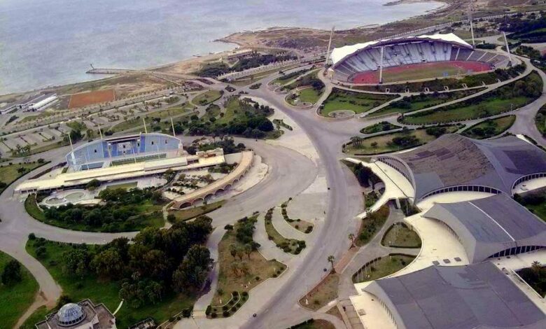 An aerial view of Latakia Sports City on the Mediterranean coast, showing stadiums, curved-roof sports halls, surrounding roads, greenery, and the sea in the background.