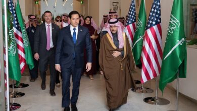Inside the Saudi Ministry of Foreign Affairs in Riyadh, officials walk through a bright corridor flanked by multiple Saudi and U.S. flags, with arched doorways and official decor visible.