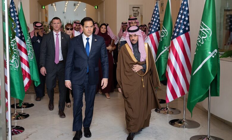 Inside the Saudi Ministry of Foreign Affairs in Riyadh, officials walk through a bright corridor flanked by multiple Saudi and U.S. flags, with arched doorways and official decor visible.