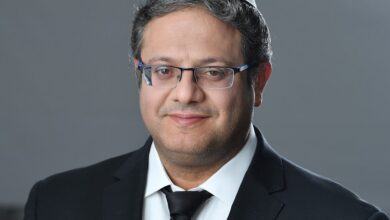 A man wearing a white kippah, dark suit, white shirt, and black tie poses against a neutral gray background, looking at the camera with a slight smile.