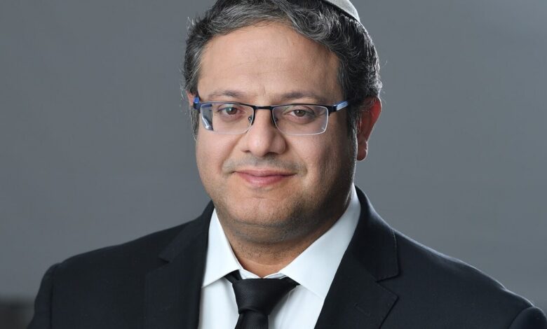 A man wearing a white kippah, dark suit, white shirt, and black tie poses against a neutral gray background, looking at the camera with a slight smile.