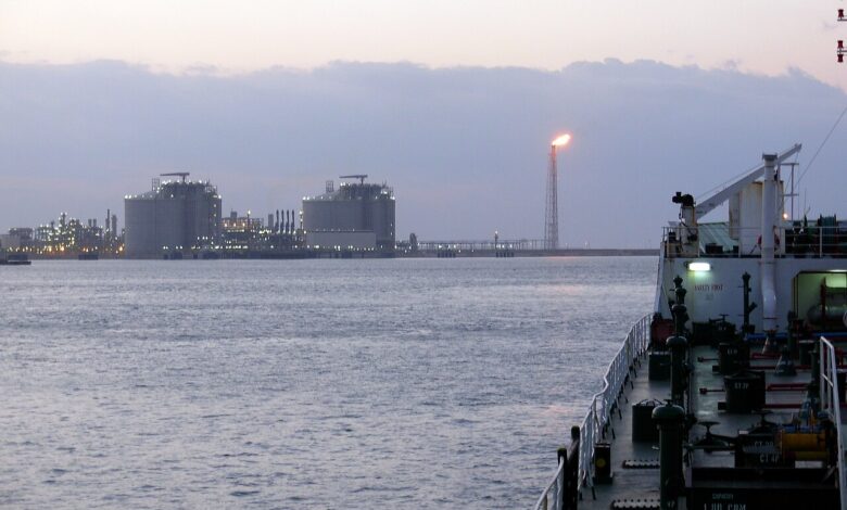 A waterside view at dusk of large LNG storage tanks and an active flare stack across a harbor, with the deck and railing of a vessel in the right foreground under a cloudy sky.