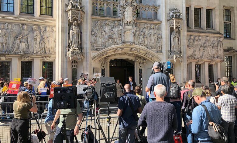 The exterior entrance of the UK Supreme Court in London with TV cameras, journalists, and onlookers gathered in front of the ornate stone facade.