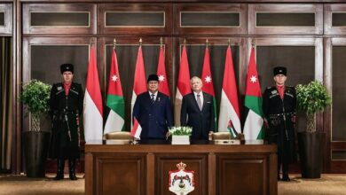 A formal signing backdrop with two officials standing before Jordanian and Indonesian flags, flanked by two Jordanian Royal Guards. A desk bearing Jordan’s national emblem and small Jordanian and Indonesian table flags is in front of them in an ornate wood-paneled government room.