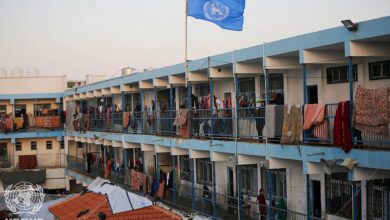A multi-storey UNRWA school building in Deir al-Balah with a blue UN flag on a pole. Blankets and clothes hang along the railings, and tents or coverings are set up in the courtyard below, indicating the school is being used as a shelter.