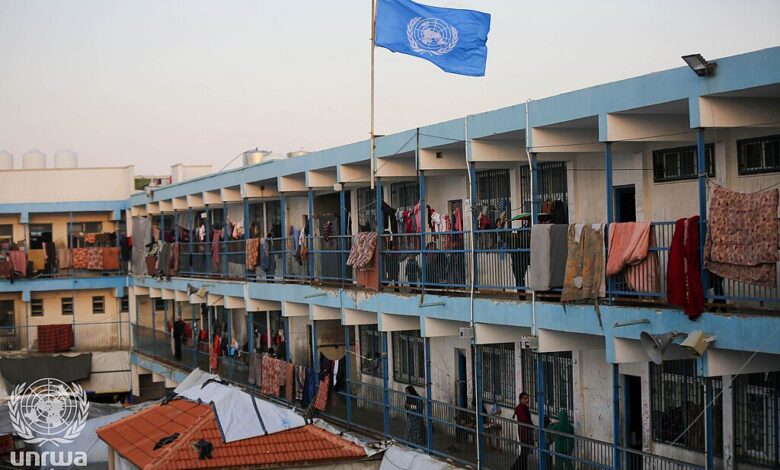 A multi-storey UNRWA school building in Deir al-Balah with a blue UN flag on a pole. Blankets and clothes hang along the railings, and tents or coverings are set up in the courtyard below, indicating the school is being used as a shelter.