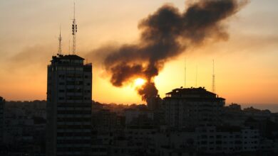 City skyline at sunset with two tall buildings in the foreground and a large plume of dark smoke rising near the setting sun over the urban area.