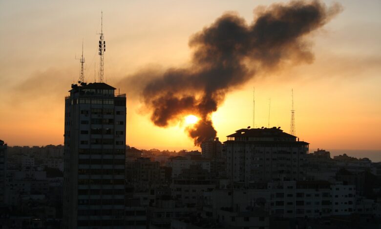 City skyline at sunset with two tall buildings in the foreground and a large plume of dark smoke rising near the setting sun over the urban area.