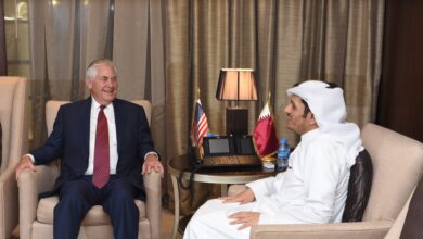 Two officials sit facing each other in armchairs, conversing. The man on the left wears a dark business suit; the man on the right wears a white thobe and headscarf. A small table between them holds a lamp and miniature U.S. and Qatari flags. Heavy curtains and a formal interior suggest an official meeting setting.