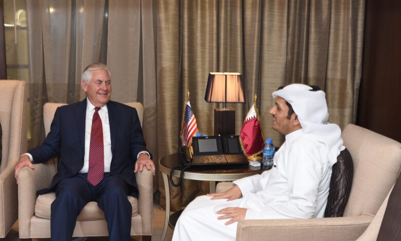 Two officials sit facing each other in armchairs, conversing. The man on the left wears a dark business suit; the man on the right wears a white thobe and headscarf. A small table between them holds a lamp and miniature U.S. and Qatari flags. Heavy curtains and a formal interior suggest an official meeting setting.