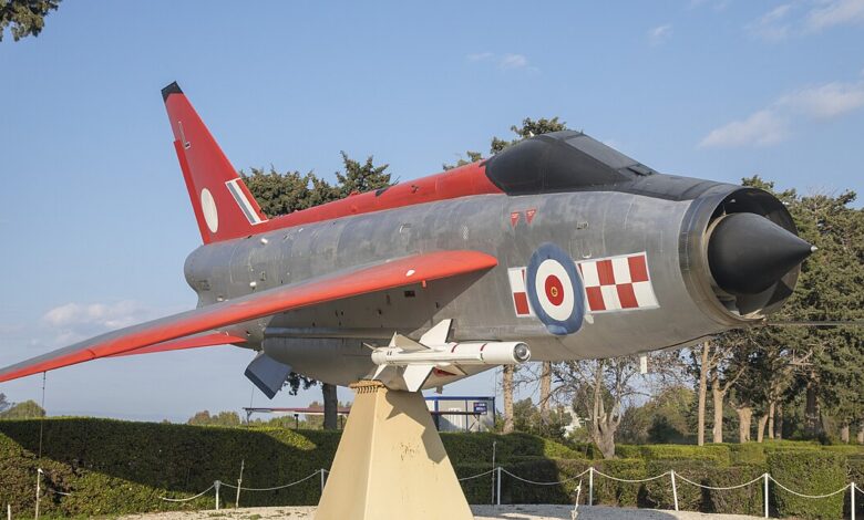 A preserved English Electric Lightning jet in RAF colors is mounted on a plinth outdoors as a gate guardian at an airbase, with low hedges, rope barriers, and a clear blue sky.