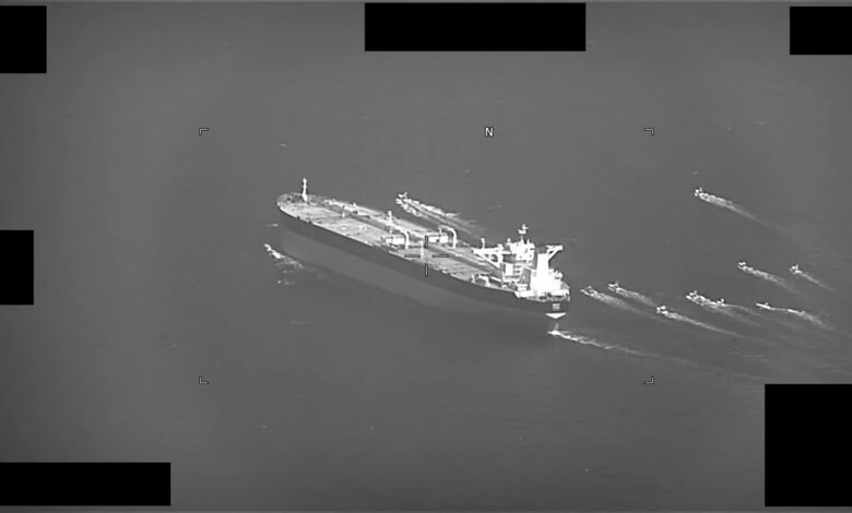 A black‐and‐white aerial view of a large oil tanker at sea accompanied by multiple small fast boats creating wakes around it.