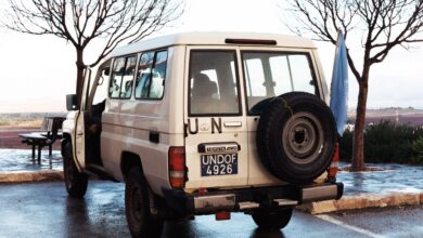 A white UN-marked Toyota Land Cruiser with UNDOF license plate and a UN flag is parked on a wet roadside viewpoint in the Golan Heights. Bare trees and a hilly landscape are visible in the background.