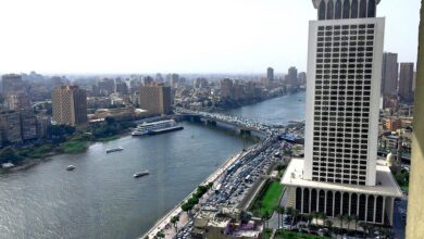 Daytime view over the Nile in Cairo with the modern Egyptian Ministry of Foreign Affairs high‐rise prominently on the right, traffic on a bridge, and the city skyline stretching into the distance under a hazy sky.