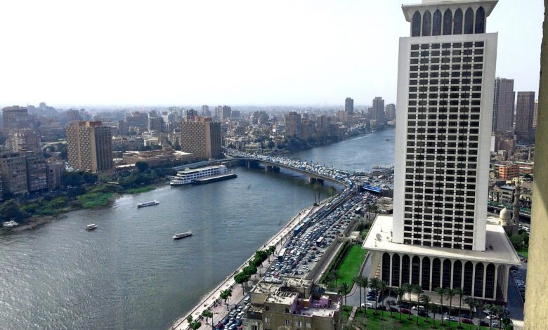 Daytime view over the Nile in Cairo with the modern Egyptian Ministry of Foreign Affairs high‐rise prominently on the right, traffic on a bridge, and the city skyline stretching into the distance under a hazy sky.