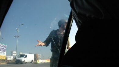 An Israeli soldier stands beside a car at a West Bank checkpoint, gesturing to the occupants. Concrete curb barriers and a passing white van are visible under a clear sky.