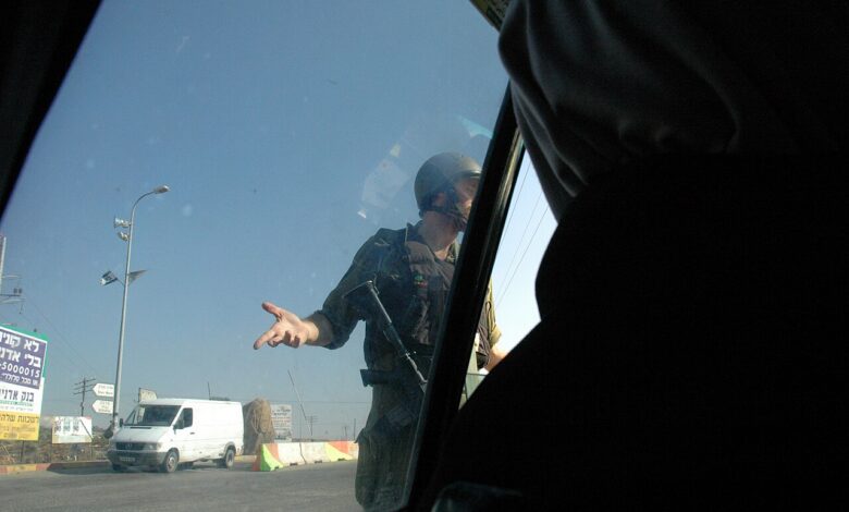 An Israeli soldier stands beside a car at a West Bank checkpoint, gesturing to the occupants. Concrete curb barriers and a passing white van are visible under a clear sky.