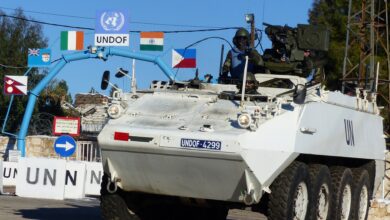 A white UN armored personnel carrier marked UNDOF-4299 drives past a gate adorned with UN and national flags at a base entrance. A helmeted peacekeeper is visible on the vehicle. Dry-stone walls and signage around the gate indicate a UNDOF facility in the Golan Heights.