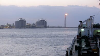 Dusk view from a ship’s deck toward a shoreline with multiple large industrial storage tanks and a tall gas flare tower burning, indicating an LNG or gas facility along the coast.