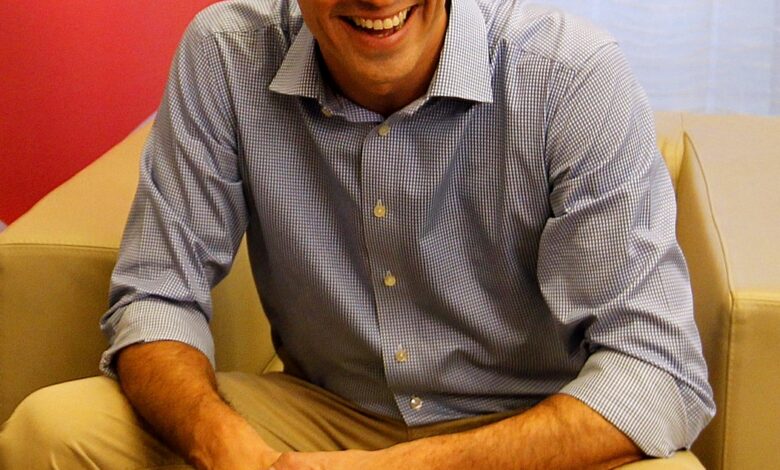 A man in a blue striped shirt and beige trousers sits on a beige armchair, smiling, with a red backdrop and soft indoor lighting.