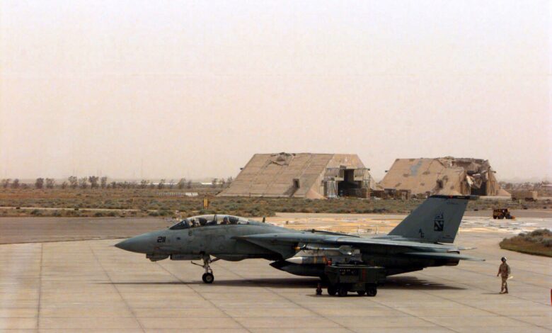 A military F-14 jet parked on a concrete ramp at an air base, with ground crew and equipment alongside and hardened aircraft shelters visible in the background under a hazy sky.