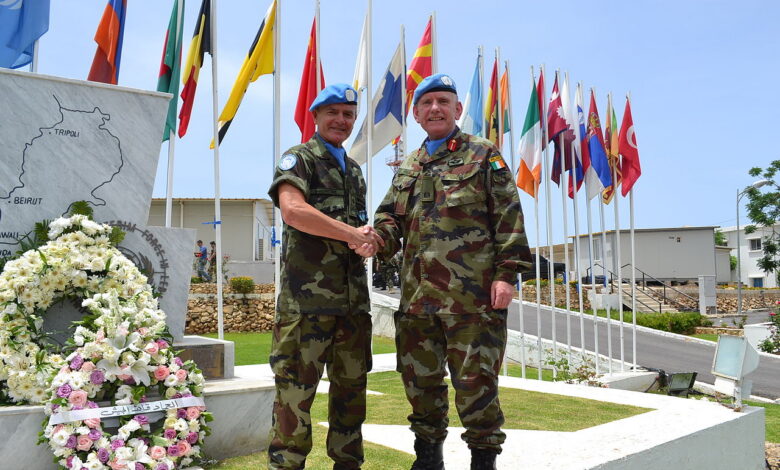 Two UN peacekeepers wearing blue berets shake hands in front of a stone monument shaped like Lebanon, with a wreath and multiple national and UN flags inside a UNIFIL compound.