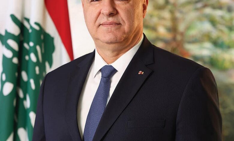 A formal portrait of a suited man standing beside a Lebanese flag, with a soft-painted backdrop of trees and sky.