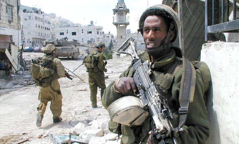 Israeli soldiers in full combat gear stand and move through a damaged urban street in Nablus, West Bank. An armored personnel carrier is visible in the background, with buildings and a mosque-like tower framing the scene.