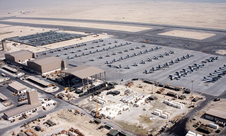 An aerial view of Shaikh Isa airfield in Bahrain with multiple rows of U.S. military aircraft parked on large aprons, hangars and support buildings, and a long runway set in a flat desert landscape.