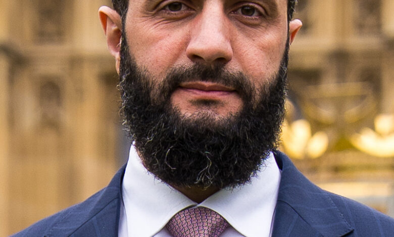 Close-up portrait of a bearded man in a navy suit and light-colored patterned tie, standing outdoors with a blurred classical stone building in the background.