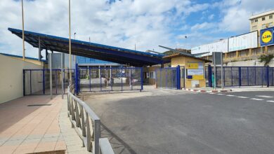 A wide view of the El Tarajal border crossing in Ceuta with blue metal gates and fencing, a covered checkpoint structure, pedestrian entry points, and a paved approach under a partly cloudy sky. A few people are visible near the gates.