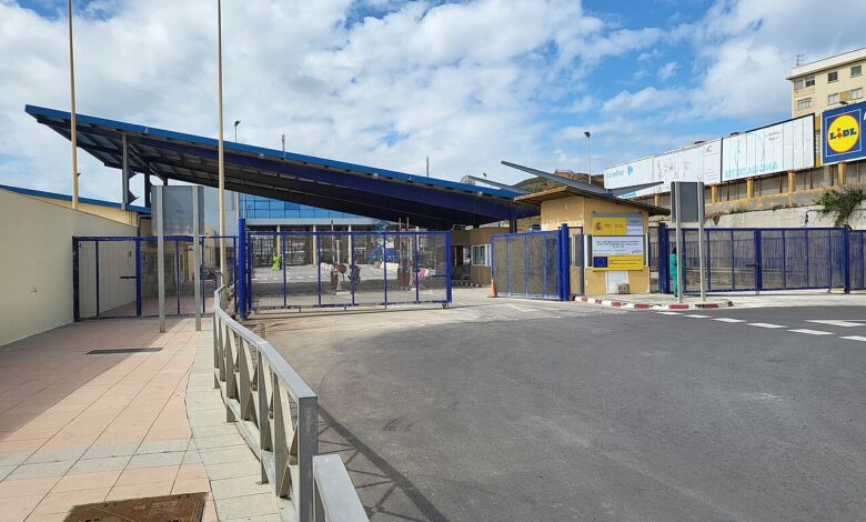 A wide view of the El Tarajal border checkpoint in Ceuta with blue metal gates and fencing, a canopy over the entry area, pedestrian pathways, and checkpoint buildings under a partly cloudy sky. A supermarket sign is visible on a wall to the right.