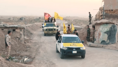 A convoy of armed Popular Mobilization Forces fighters in pickup trucks with yellow PMF flags moves along a dirt road in a rural Iraqi landscape, with additional fighters standing near a small outpost and mural-painted wall.