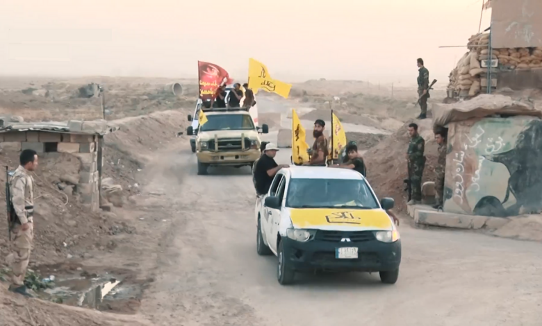 A convoy of armed Popular Mobilization Forces fighters in pickup trucks with yellow PMF flags moves along a dirt road in a rural Iraqi landscape, with additional fighters standing near a small outpost and mural-painted wall.