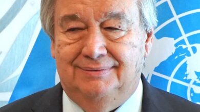 A mature man in a suit and red tie stands before a blue United Nations flag, photographed head-and-shoulders and facing the camera with a slight smile.