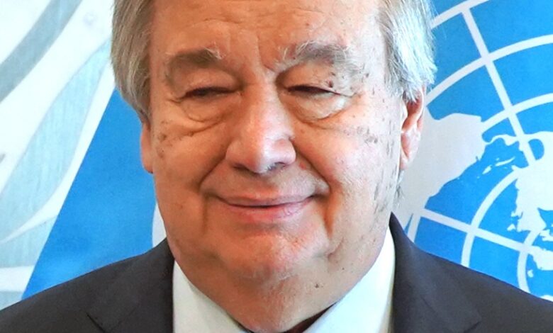 A mature man in a suit and red tie stands before a blue United Nations flag, photographed head-and-shoulders and facing the camera with a slight smile.