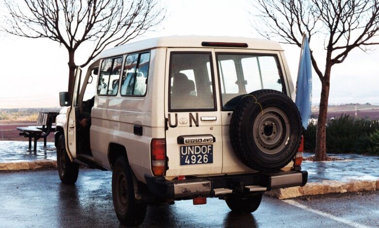 A white UN Toyota Land Cruiser with an UNDOF license plate and UN flag is parked on a roadside overlooking fields in the Golan Heights. Bare trees and wet pavement frame the vehicle.