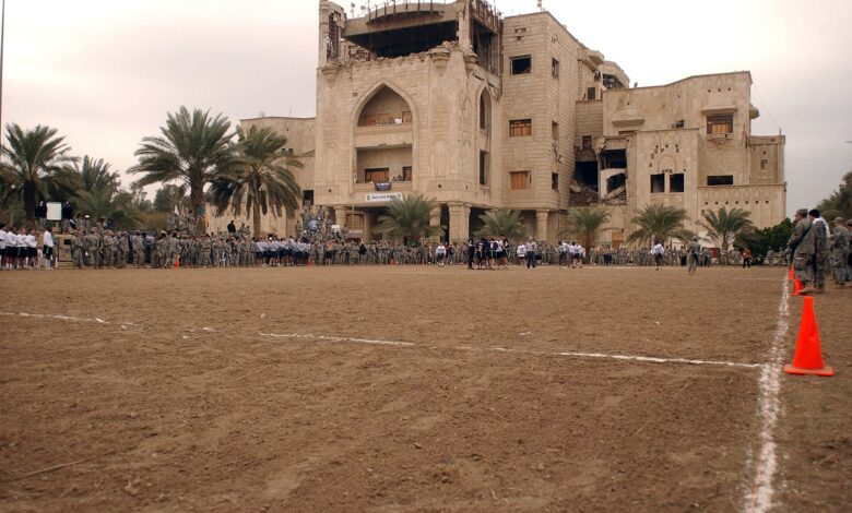 A wide exterior view of Al Salam Palace in Baghdad with palm trees and a large sandy field in the foreground. Military personnel and civilians line the edges of the field, with orange cones marking boundaries, and the multi-story palace building rises behind them.