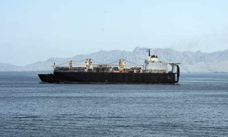 A dark-hulled dry cargo ship sails across calm blue waters with rugged, arid mountains lining the distant shore under a clear sky in the Strait of Hormuz.